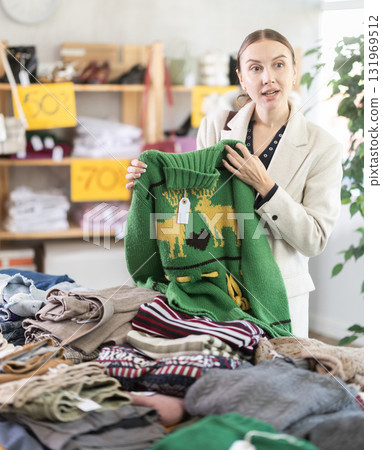European female buyer standing in clothing market and carefully examines selection of sweaters on Black Friday sale of winter clothing collection European female buyer standing in clothing market and carefully examines selection of sweaters on Black Friday sale of winter clothing collection 131969512