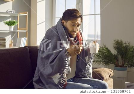 Sick young man checking his temperature wrapped in a blanket at home. 131969761