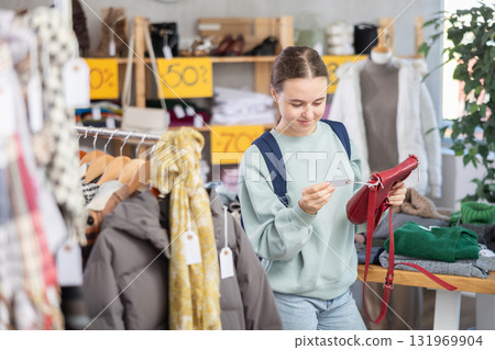 Happy teen girl standing in interior of clothing boutique and choosing fashionable handbag Happy teen girl standing in interior of clothing boutique and choosing fashionable handbag 131969904