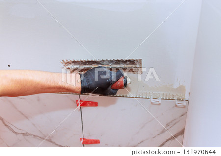 Construction worker uses trowel to spread tile adhesive on wall during bathroom remodel 131970644