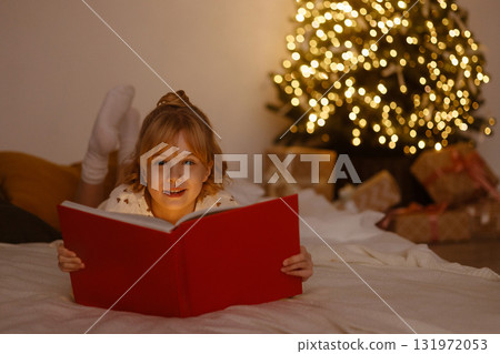 Little girl lying on bed reading red book near Christmas tree with lights and gifts 131972053