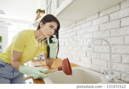 Worried young woman in gloves holding plunger and calling plumber while trying to fix clogged sink. Worried young woman in gloves holding plunger and calling plumber while trying to fix clogged sink. 131972522
