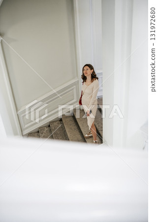 A fashionable woman in a beige dress stands on the staircase, elegantly looking upwards in a well-lit interior with neutral decor and classic architecture. A fashionable woman in a beige dress stands on the staircase, elegantly looking upwards in a well-lit interior with neutral decor and classic architecture. 131972680