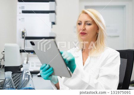 Blonde woman without mask wearing gloves recording laboratory experiment results on a tablet while standing in a chemistry lab with flasks, beakers, and HPLC equipment Blonde woman without mask wearing gloves recording laboratory experiment results on a tablet while standing in a chemistry lab with flasks, beakers, and HPLC equipment 131975091