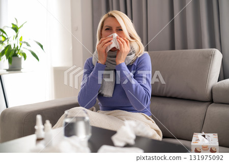 Middle aged blonde woman sitting on couch at home, blowing her nose into a tissue. Medicines and tissues are placed on the table next to her. Concept of cold, flu, covid, home treatment, and recovery. 131975092