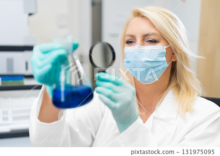 Middle aged female scientist wearing mask in lab, studying chemical solution in flask through magnifying glass. Middle aged female scientist wearing mask in lab, studying chemical solution in flask through magnifying glass. 131975095