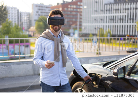 A man wearing a VR headset charges his electric vehicle while holding a smartphone, showcasing modern technology and sustainable transportation. 131975305