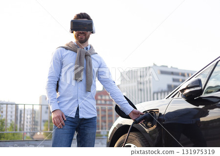 A man wearing a VR headset charges an electric vehicle, showcasing the future of technology and sustainable transportation. A man wearing a VR headset charges an electric vehicle, showcasing the future of technology and sustainable transportation. 131975319