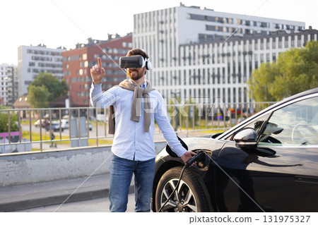 A man wearing a VR headset points while charging his electric vehicle in an urban setting. 131975327