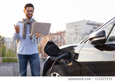 A man stands next to his electric vehicle while it charges, holding a laptop and appearing to be working or video conferencing. A man stands next to his electric vehicle while it charges, holding a laptop and appearing to be working or video conferencing. 131975353