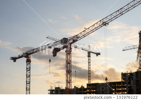 Cranes loom at a construction site against a colorful sunset, as workers engage in various tasks to progress the building project. Cranes loom at a construction site against a colorful sunset, as workers engage in various tasks to progress the building project. 131976122