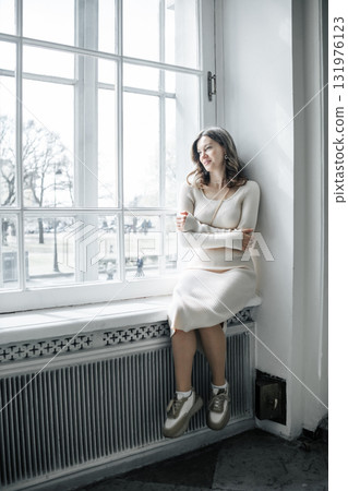 A woman in a cozy light sweater relaxes by a large window, gazing outside at the sunny scenery, showcasing a peaceful moment of reflection. 131976123