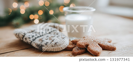 Christmas mitten, gingerbread cookie, and glass of milk rest on wooden table with festive lights in background, creating cozy holiday atmosphere and warm seasonal feeling 131977341