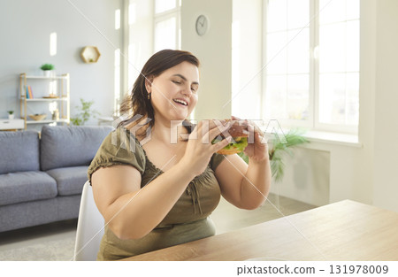 Young fat woman sitting at the table at home holding tasty burger in hands. 131978009