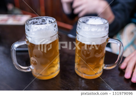 Prague, Czech Republic, August 7, 2023. Two mugs of beer side by side. In the blurred background, the hands of those enjoying them. A lifestyle of shared drinking. Prague, Czech Republic, August 7, 2023. Two mugs of beer side by side. In the blurred background, the hands of those enjoying them. A lifestyle of shared drinking. 131979096