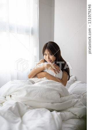 Smiling young woman hugging a pillow in bed while basking in the morning sun Smiling young woman hugging a pillow in bed while basking in the morning sun 131979246