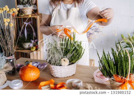 Woman creating a floral gift basket with orange and white ribbon on a wooden table. Thanksgiving aesthetic, autumn DIY, seasonal floristry, cozy living trends Woman creating a floral gift basket with orange and white ribbon on a wooden table. Thanksgiving aesthetic, autumn DIY, seasonal floristry, cozy living trends 131980152
