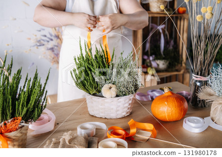 Woman arranging white heather and evergreen branches into a decorative basket on a craft table. Thanksgiving workshop, seasonal DIY, natural decor, creative wellness 131980178