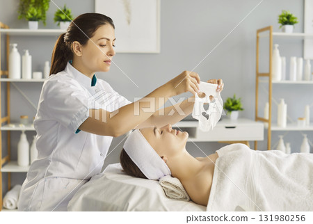 Female beautician applying moisturizing sheet mask on young woman face in beauty spa salon. Female beautician applying moisturizing sheet mask on young woman face in beauty spa salon. 131980256