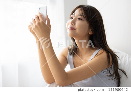 一位年輕女子躺在床上,一邊操作智慧型手機,一邊沐浴在窗外清晨的陽光 一位年輕女子躺在床上,一邊操作智慧型手機,一邊沐浴在窗外清晨的陽光 131980642
