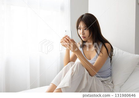 A young woman operating a smartphone on her bed while basking in the morning sun from the window 131980646