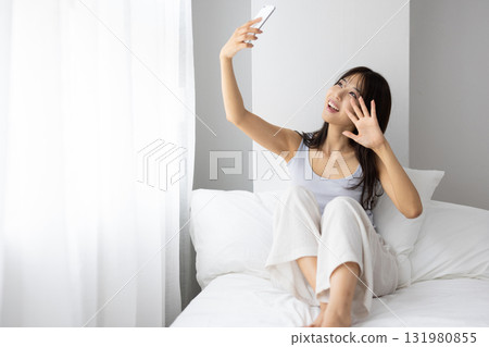 一名年輕女子躺在床上，一邊沐浴在窗外的晨光，一邊用智慧型手機進行視訊通話。 131980855