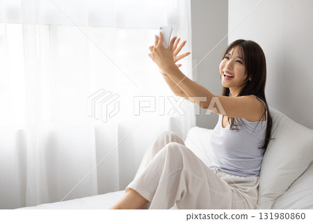 A young woman making a video call on her smartphone while basking in the morning sun from the window on her bed. 131980860