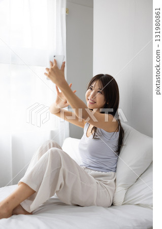 A young woman making a video call on her smartphone while basking in the morning sun from the window on her bed. 131980861