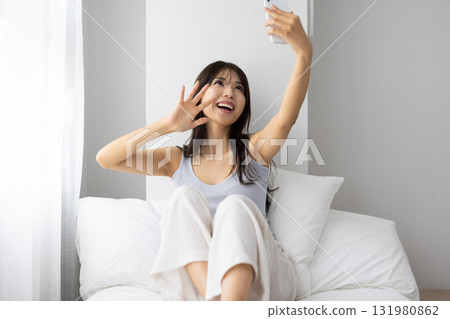 A young woman making a video call on her smartphone while basking in the morning sun from the window on her bed. 131980862