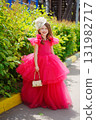 A girl in a lush pink dress, hat and with a golden handbag near the green bushes 131982717