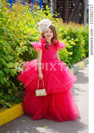 A girl in a lush pink dress, hat and with a golden handbag near the green bushes 131982717