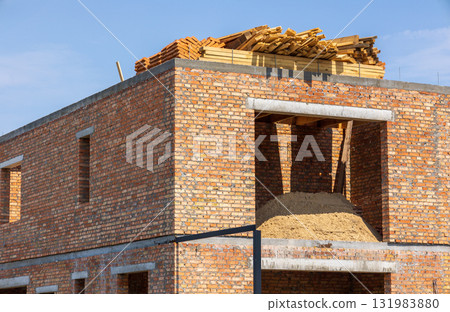 A residential building is in development with brick walls, a pile of sand, and wooden beams stacked on the roof under a clear sky 131983880