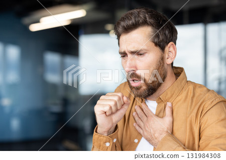 Adult man coughing into his fist, clutching his chest and looking unwell from a cold or respiratory illness, showing discomfort and fatigue in a studio setting 131984308