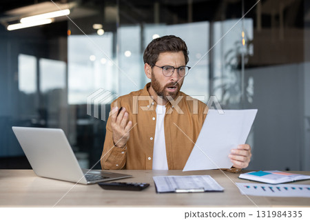 Stressed man in optical glasses sitting at office desk, holding paperwork, and reacting with a shocked and confused expression after seeing a bill or unexpected debt Stressed man in optical glasses sitting at office desk, holding paperwork, and reacting with a shocked and confused expression after seeing a bill or unexpected debt 131984335