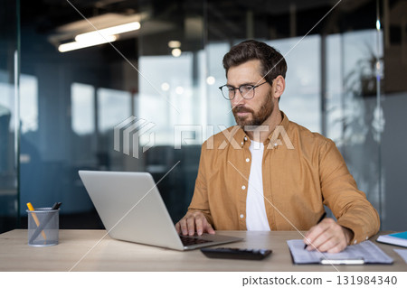 Businessman in glasses and a brown shirt is actively working at his office desk, typing on a laptop and writing notes, demonstrating focus on work, productivity, and professional multitasking Businessman in glasses and a brown shirt is actively working at his office desk, typing on a laptop and writing notes, demonstrating focus on work, productivity, and professional multitasking 131984340