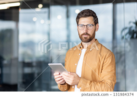 Confident bearded man with glasses standing in a modern office, holding a tablet and engaging with digital technology for work, communication and business planning Confident bearded man with glasses standing in a modern office, holding a tablet and engaging with digital technology for work, communication and business planning 131984374
