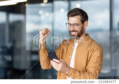Enthusiastic man smiling widely and raising his fist in an office environment, celebrating good news received on his mobile phone, achieving a business goal or personal success Enthusiastic man smiling widely and raising his fist in an office environment, celebrating good news received on his mobile phone, achieving a business goal or personal success 131984377