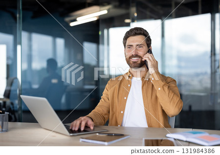 Businessman on a phone call smiling while typing on a laptop at a modern glass-partitioned desk, multitasking in a bright office, confident and productive during the workday 131984386