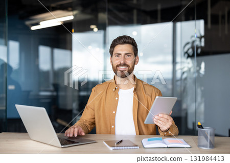 Man smiling at the camera, confidently working on a laptop and holding a digital tablet at an office desk, representing modern business communication and success Man smiling at the camera, confidently working on a laptop and holding a digital tablet at an office desk, representing modern business communication and success 131984413