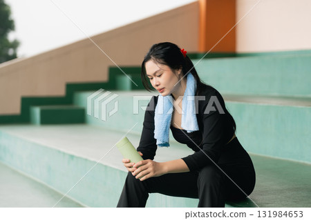 Young woman taking a break on stadium steps, holding a green drink, wearing casual sportswear with a towel around her neck and reflecting on her workout routine 131984653