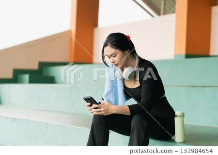 Young woman in athletic wear sitting on bleachers with earphones, towel, and phone, focused on mobile device during break after workout session 131984654