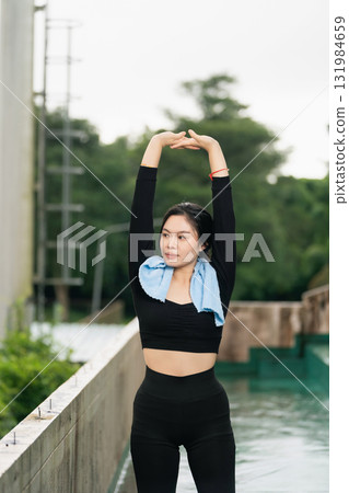 Young woman practicing yoga poses outdoors near a water body while wearing athletic attire and a blue towel around her neck for fitness exercise routine Young woman practicing yoga poses outdoors near a water body while wearing athletic attire and a blue towel around her neck for fitness exercise routine 131984659