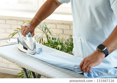 Man ironing blue shirt on board in bright indoor space during the day Man ironing blue shirt on board in bright indoor space during the day 131987116