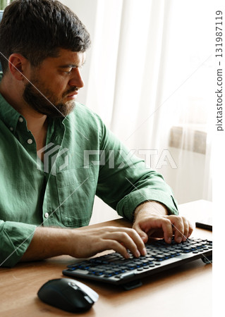 Young man typing on keyboard while sitting at wooden desk in bright room during daytime 131987119
