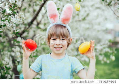Easter Egg Hunt with Child Carrying Bright Plastic Eggs. Smiling child wearing bunny ears holding a wicker basket filled with colorful Easter eggs in a blooming spring garden 131987594