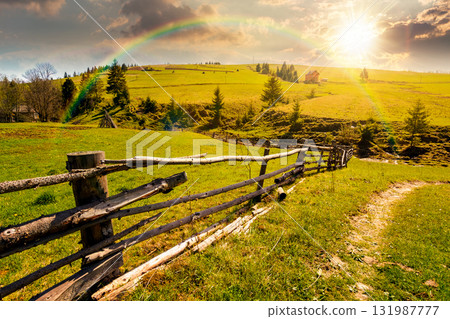 beautiful rural landscape in mountains at sunset in spring. road near the wooden fence through the grassy pasture with haystack and coniferous trees on hillside in evening light beautiful rural landscape in mountains at sunset in spring. road near the wooden fence through the grassy pasture with haystack and coniferous trees on hillside in evening light 131987777
