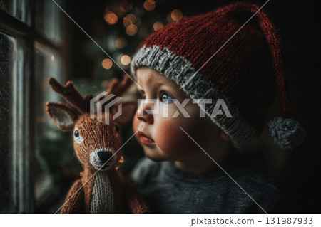 A toddler wearing a Santa hat looks in awe at the snowy window. Holding a plush reindeer, the child enjoys the winter scenery on a cold day, lost in imagination and joy 131987933