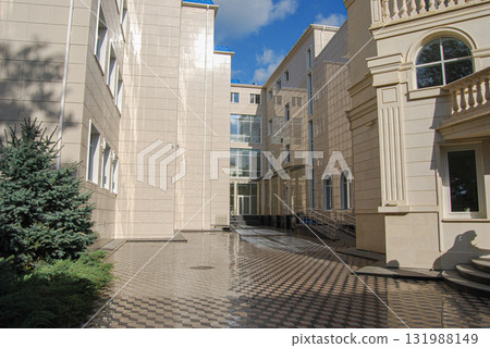 Modern building exterior with landscaping and tiled pavement. Modern building with a beige tiled facade and multiple white-framed windows. Modern building exterior with landscaping and tiled pavement. Modern building with a beige tiled facade and multiple white-framed windows. 131988149