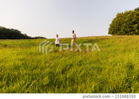 A father and his son walk happily through a vibrant green meadow on a sunny afternoon. They are exploring nature, enjoying quality time together amidst the beautiful scenery 131988193