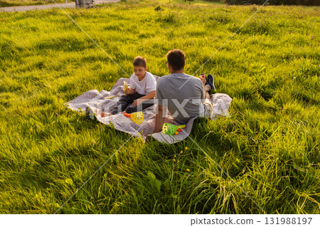 A father and his young son share a relaxing picnic on a soft blanket in a vibrant green field. They enjoy snacks and each other's company under the warm sun 131988197